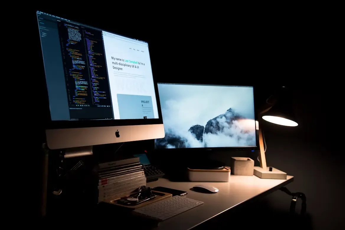 A dark home studio desk with a monitor showing code and a website design